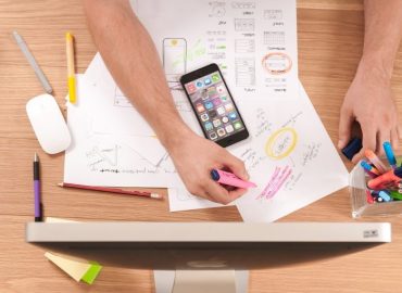 Person highlighting notes on papers at a desk with pens, markers, a smartphone, and a computer monitor.