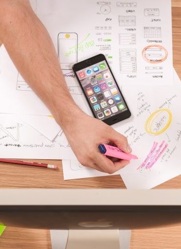 Person highlighting notes on papers at a desk with pens, markers, a smartphone, and a computer monitor.