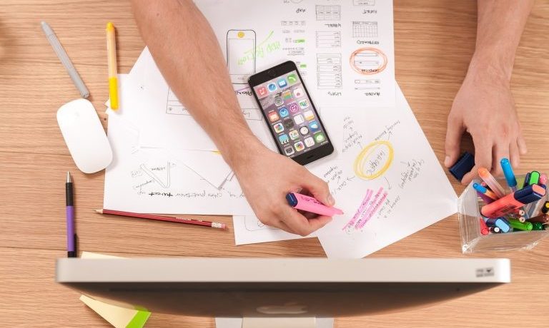 Person highlighting notes on papers at a desk with pens, markers, a smartphone, and a computer monitor.